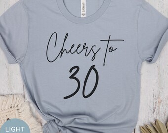 May include: Pale blue t-shirt featuring the black text "Cheers to 30". The number 30 is in a bold font. The shirt has rolled-up sleeves and is made of a soft material. The t-shirt is a light blue colour.