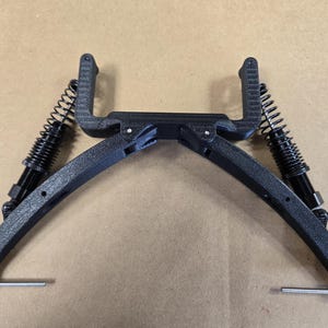 May include: A black, mechanical device with a curved base and two vertical supports connected by a horizontal bar. It features two black springs and metal pins. The device is designed for a specific function, likely related to clamping or holding.