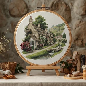 May include: A framed embroidery hoop featuring a detailed landscape scene of quaint cottages along a stream. The artwork is displayed on a small wooden easel. The scene includes lush greenery, a small bridge, and a stone wall background.