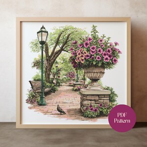 May include: A framed cross-stitch pattern depicting a park scene. The image shows a brick pathway, benches, and a large stone planter filled with pink flowers. A green lamppost and trees line the path. A bird stands on the path. The image has a PDF Pattern label.