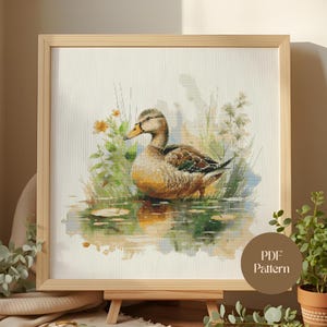 Puede incluir: Un patrón de punto de cruz enmarcado de un pato en un estanque. El pato tiene plumas marrones, beige y blancas, un pico amarillo y está rodeado de plantas verdes y flores amarillas. La obra de arte está en un marco de madera clara.