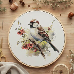 Puede incluir: Bordado de punto de cruz de un pájaro en una rama con flores rojas, en un bastidor de madera. El pájaro tiene plumas marrones, blancas y grises. Suministros de costura rodean el bastidor.