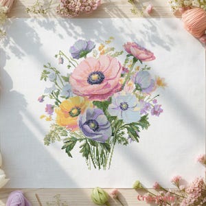 Puede incluir: Un diseño floral de punto de cruz con un ramo de flores coloridas. El ramo incluye flores rosas, amarillas y moradas con follaje verde. El diseño está sobre un fondo blanco, rodeado de materiales de manualidades.