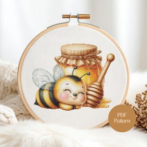 May include: Cross-stitch embroidery hoop with a cartoon bee, honey jar, and dipper. The bee is yellow and black with a smiling face. The honey jar is filled with golden honey. The text "PDF Pattern" is on the bottom right.