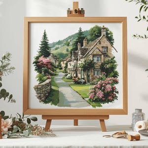 May include: A framed watercolor print of a quaint village scene. The artwork depicts stone houses with green foliage, a winding road, and a stone wall. Pink flowers and a tall evergreen tree add color to the landscape. The print is displayed on a wooden easel.