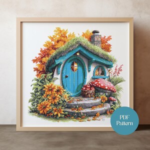 Peut inclure: Une broderie au point de croix encadrée représentant une maisonnette fantaisiste avec une porte bleue et un toit vert. La scène est ornée de feuillage d'automne orange et jaune, d'un champignon rouge et d'un panier de pommes. Le texte "PDF Pattern" est sur un cercle bleu.