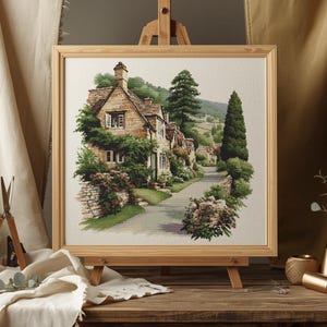 May include: A framed watercolor painting of a quaint village scene. The artwork depicts stone houses with green foliage, a winding road, and tall trees. The painting is displayed on a wooden easel, with a neutral-toned background.