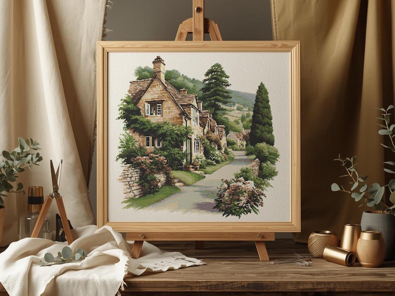 May include: A framed watercolor painting of a quaint village scene. The artwork depicts stone houses with green foliage, a winding road, and tall trees. The painting is displayed on a wooden easel, with a neutral-toned background.