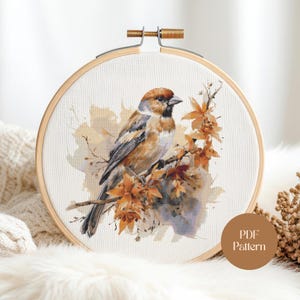 Puede incluir: Un bastidor de bordado de punto de cruz con un pájaro en una rama con hojas de otoño. El pájaro tiene plumas marrones, grises y blancas. El bordado está en un bastidor de madera, con el texto "PDF Pattern".