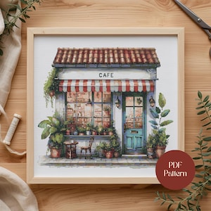 May include: Cross-stitch pattern of a quaint cafe with a red and white striped awning, a teal door, and the word "CAFE" above the entrance. The scene is framed by a wooden border and includes potted plants and a small table.