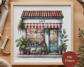 May include: Cross-stitch pattern of a quaint cafe with a red and white striped awning, a teal door, and the word "CAFE" above the entrance. The scene is framed by a wooden border and includes potted plants and a small table.