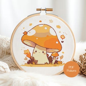 Può includere: Un telaio da ricamo a punto croce con un design a fungo a cartoni animati. Il fungo ha un gambo color crema e un cappello arancione brillante con punti bianchi. Il testo "PDF Pattern" è su un adesivo circolare.