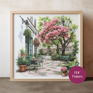 May include: Framed cross-stitch of a European cafe scene. The artwork features a cafe with green doors and tables, a large tree with pink blossoms, and potted plants. The words "PDF Pattern" are on a purple circle in the lower right corner.