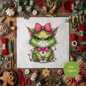 Op de afbeelding: Kruissteekpatroon van een groene kat met een boze blik, roze strik en vlinderdas, en kerstverlichting. Het ontwerp staat op witte stof, omringd door knutselbenodigdheden.