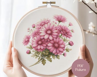 May include: A cross-stitch embroidery of pink daisies in a wooden hoop. The floral design features various shades of pink and green. The text "PDF Pattern" is on a pink circle in the lower right corner.