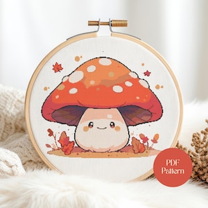 May include: A cross-stitch mushroom design in a wooden embroidery hoop. The mushroom has a red-orange cap with white spots and a smiling face. Autumn leaves and the text "PDF Pattern" are also visible.