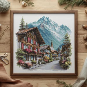 Puede incluir: Un bordado enmarcado que representa un pueblo europeo al pie de una montaña nevada. Los edificios tienen tejados marrones y contraventanas rojas, con flores de colores en los balcones. La obra de arte está en un marco de madera marrón.