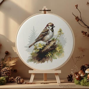 Könnte beinhalten: Gerahmte Kreuzstich-Stickerei eines Spatzen auf einem Ast, vor einer Waldkulisse. Das Kunstwerk wird auf einer Holzstaffelei präsentiert, umgeben von natürlichen Elementen wie Tannenzapfen und Pilzen.