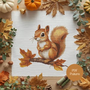 Könnte beinhalten: Ein Kreuzstichmuster mit einem Eichhörnchen auf einem Ast mit Herbstblättern. Das Eichhörnchen ist braun mit einem flauschigen Schwanz. Das Muster ist von Herbstlaub und Kürbissen umgeben. Der Text "PDF Pattern" befindet sich in einem Kreis.