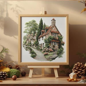 May include: A framed watercolor painting of a quaint cottage scene. The artwork depicts a stone house with a red-tiled roof, surrounded by lush greenery and a winding pathway. The painting is displayed on a small wooden easel, with autumn-themed decorations.
