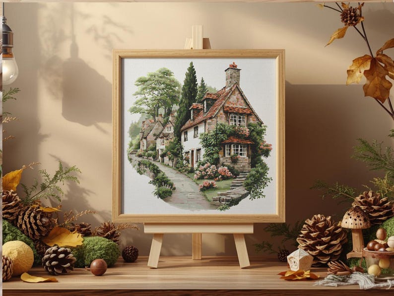 May include: A framed watercolor painting of a quaint cottage scene. The artwork depicts a stone house with a red-tiled roof, surrounded by lush greenery and a winding pathway. The painting is displayed on a small wooden easel, with autumn-themed decorations.