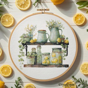 May include: A cross-stitch embroidery hoop featuring a watercolor design of a shelf with jars, pitchers, and plants, with lemons scattered around. The embroidery hoop is surrounded by fresh lemons and greenery.