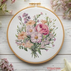 May include: A completed cross-stitch embroidery of a floral bouquet in a wooden hoop. The bouquet features pink, peach, and purple flowers with green foliage. The hoop is made of light brown wood. The text "CrossCount Studio" is visible at the bottom.