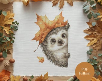May include: A cross-stitch pattern featuring a charming hedgehog peeking out from behind a white canvas, wearing a large orange maple leaf. The design is surrounded by autumn leaves, pumpkins, and the text "PDF Pattern".