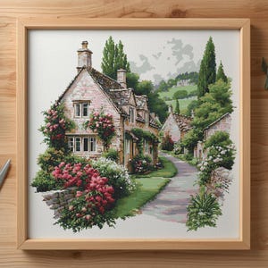 May include: A framed cross-stitch of a quaint village scene. The artwork depicts stone cottages with climbing roses and a winding road. The color palette includes greens, pinks, and beige, creating a serene, pastoral feel. The frame is made of light-colored wood.