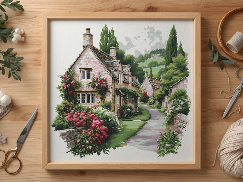 May include: A framed cross-stitch of a quaint village scene. The artwork depicts stone cottages with climbing roses and a winding road. The color palette includes greens, pinks, and beige, creating a serene, pastoral feel. The frame is made of light-colored wood.