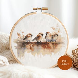 May include: A cross-stitch embroidery featuring four small birds perched on a branch. The birds have brown and tan feathers, set against a watercolor-style background in warm, earthy tones. The design is framed in a wooden embroidery hoop. The text "PDF Pattern" is visible.