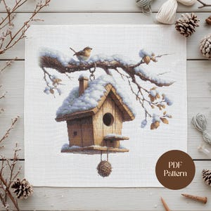 Op de afbeelding: Een kruissteekpatroon met een houten vogelhuisje bedekt met sneeuw, hangend aan een met sneeuw bedekte tak. Een kleine vogel zit op de tak. Het vogelhuisje heeft een ronde ingang en een kleine zitstok. De tekst "PDF Pattern" staat in een cirkel.