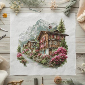 Puede incluir: Patrón de punto de cruz de un pueblo de montaña con casas de madera, contraventanas rojas y flores rosas. Un teleférico asciende una montaña. Suministros de costura rodean la escena.