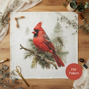 Op de afbeelding: Een kruissteekpatroon met een felrode kardinaalvogel op een tak, tegen een achtergrond van groenblijvende bomen. Het ontwerp staat op witte stof, met de woorden "PDF Pattern" in een cirkelvormig ontwerp.