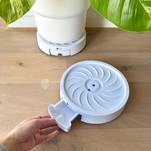 May include: White plastic plant pot saucer with a removable tray. The saucer has a spiral design and a small logo. A hand is shown pulling out the tray. The saucer is designed to catch excess water from a potted plant.