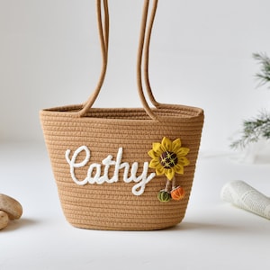 May include: A woven brown tote bag with the name "Cathy" embroidered in white. A yellow sunflower and small decorative accents adorn the front. The bag has two rope handles and is set against a white background.