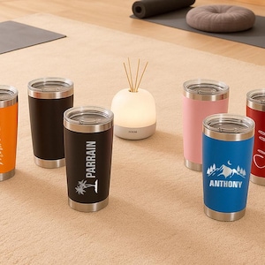 May include: A collection of insulated tumblers in various colors, including orange, black, pink, blue, and red. Each tumbler features a silver lid and personalized text or graphics. The tumblers are displayed on a neutral-toned rug.