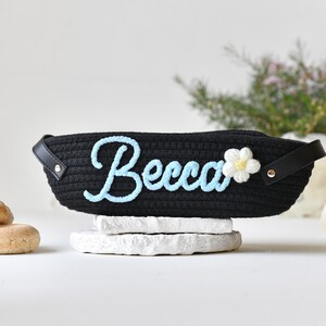 Op de afbeelding: Zwarte geweven mand met een blauwe geborduurde naam "Becca" en een witte bloem met een gele kern.