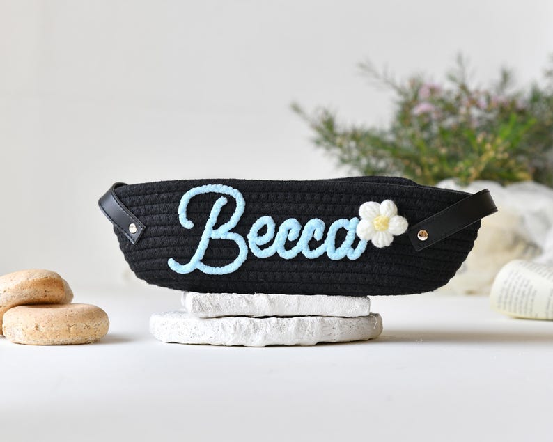 Op de afbeelding: Zwarte geweven mand met een blauwe geborduurde naam "Becca" en een witte bloem met een gele kern.