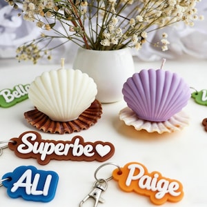 K&ouml;nnte beinhalten: Ein Stillleben mit zwei muschelf&ouml;rmigen Kerzen, eine wei&szlig;e und eine lila, auf dekorativen Muscheln. Schl&uuml;sselanh&auml;nger mit Namen wie "Superbe" und "Paige" sind verstreut. Eine kleine wei&szlig;e Vase enth&auml;lt Trockenblumen.