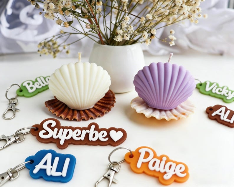 K&ouml;nnte beinhalten: Ein Stillleben mit zwei muschelf&ouml;rmigen Kerzen, eine wei&szlig;e und eine lila, auf dekorativen Muscheln. Schl&uuml;sselanh&auml;nger mit Namen wie "Superbe" und "Paige" sind verstreut. Eine kleine wei&szlig;e Vase enth&auml;lt Trockenblumen.