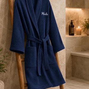 May include: A dark navy blue waffle-knit bathrobe featuring a shawl collar, long sleeves, and a matching tie belt. The left chest area is embroidered with the name Marlie in a white cursive script. The robe has two front patch pockets.