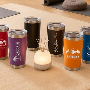 May include: A collection of insulated tumblers in various colors, including red, purple, black, orange, and blue. Each tumbler has a silver lid and base. Some tumblers feature personalized text and graphics, such as names and dates, making them ideal for gifts.