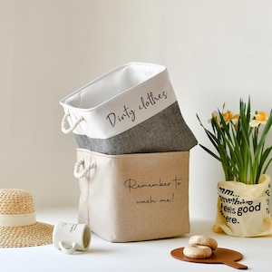 May include: Two canvas laundry baskets with rope handles. The top basket is white with grey trim and the text "Dirty clothes". The bottom basket is beige with the text "Remember to wash me!"