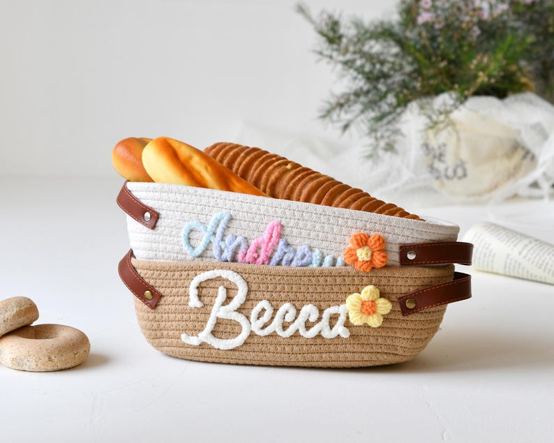 Op de afbeelding: Twee geweven manden in wit en bruin met bruine leren handvatten. De bovenste mand heeft de naam "Andrew" geborduurd in roze en blauwe draad. De onderste mand heeft de naam "Becca" geborduurd in witte draad. Beide manden hebben een klein bloemetje geborduurd aan de zijkant. De manden zijn gevuld met brood.