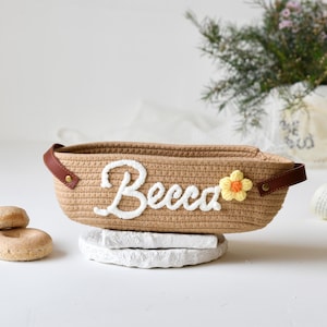 Op de afbeelding: Een bruine geweven mand met een witte geborduurde naam "Becca" en een gele gehaakte bloem. De mand staat op een wit oppervlak.