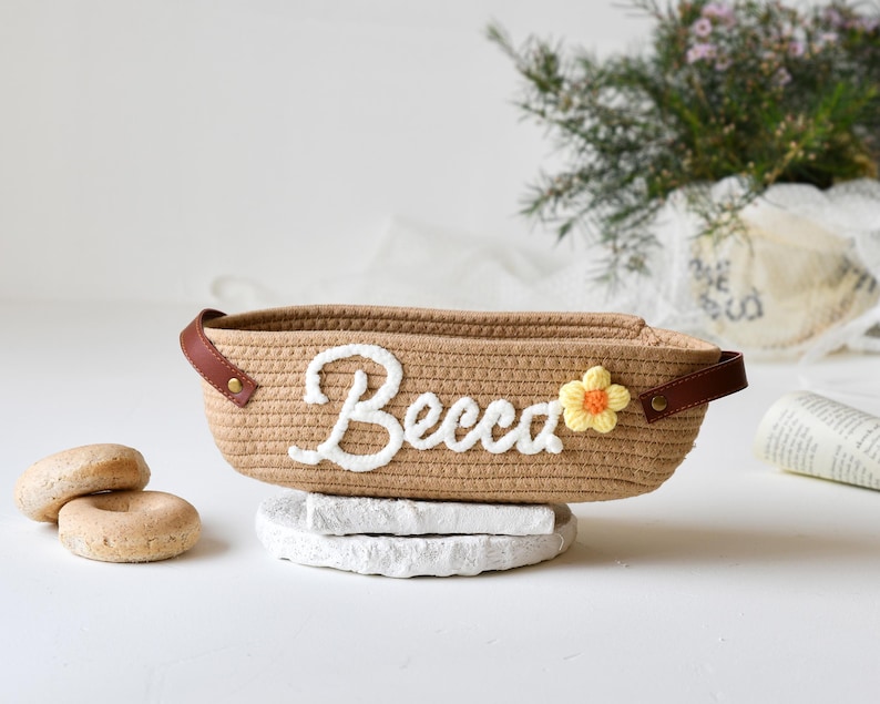 Op de afbeelding: Een bruine geweven mand met een witte geborduurde naam "Becca" en een gele gehaakte bloem. De mand staat op een wit oppervlak.