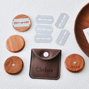 Puede incluir: Un conjunto de portacuchillas y cuchillas de afeitar de madera, con una funda de cuero marrón con la etiqueta "Chelsea". Los soportes de madera redondos tienen nombres grabados. Las cuchillas plateadas están dispuestas cerca de los soportes.