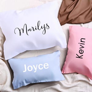 May include: Four personalised pillows in white, light blue, and pink. The white pillow has the name "Marilys" in black script. The light blue pillow has the name "Joyce" in white block letters. The pink pillow has the name "Kevin" in black block letters.