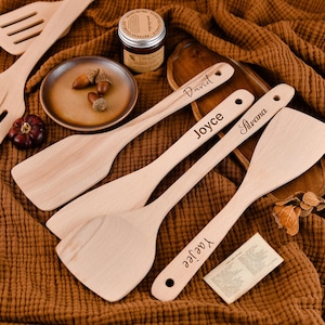 Könnte beinhalten: Eine Sammlung von Küchenutensilien aus Holz, darunter Spatel und Schöpflöffel, mit personalisierten Namen auf den Griffen. Die Utensilien sind auf einer braunen Oberfläche angeordnet, zusammen mit einem kleinen Teller mit Eicheln und einem Glas Konfitüre.